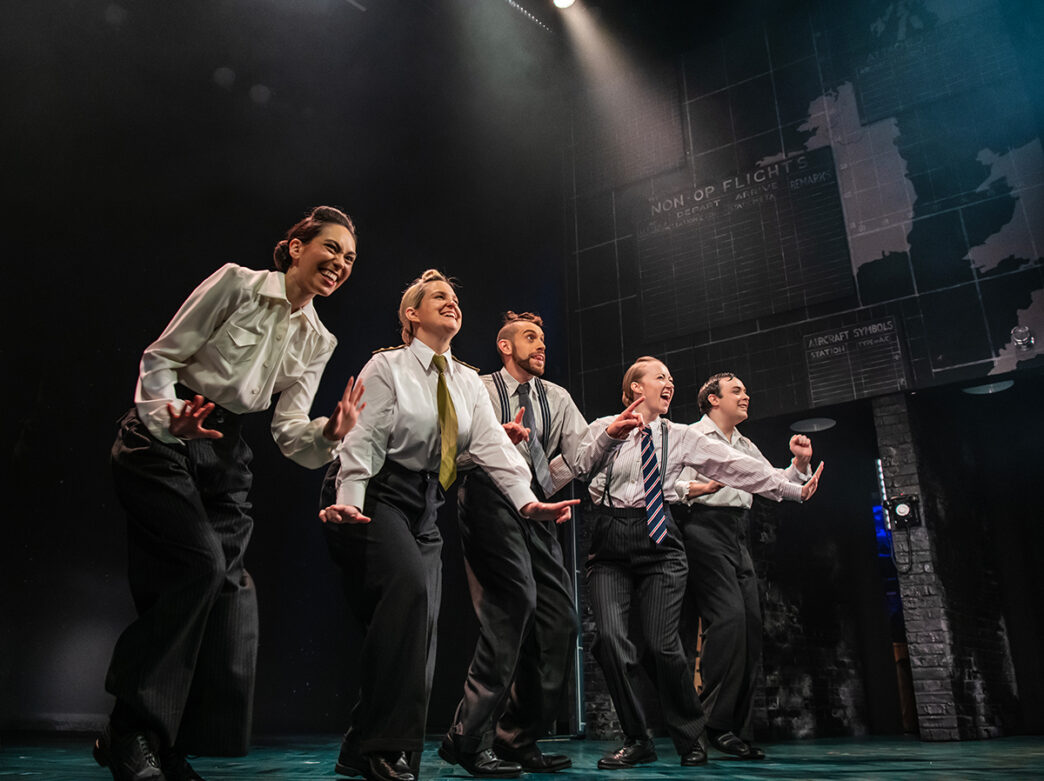 Five performers in shirts, ties and trousers sing and dance together on a dimly lit stage.