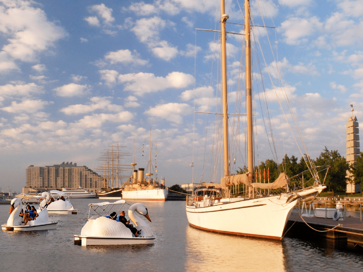 Visitors pedal white swan-shaped boats on the Delaware River waterfront, with sailboats and historic ships docked at the shoreline.