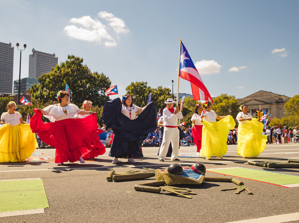 Bailarines con trajes vaporosos de color rojo, blanco y amarillo interpretan una danza tradicional a lo largo de la avenida Benjamin Franklin Parkway, mientras un participante porta una gran bandera puertorriqueña en alto.