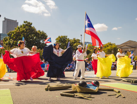 Bailarines con trajes vaporosos de color rojo, blanco y amarillo interpretan una danza tradicional a lo largo de la avenida Benjamin Franklin Parkway, mientras un participante porta una gran bandera puertorriqueña en alto.