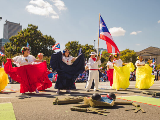 Dancers in flowing red, white and yellow costumes perform a traditional dance along the Benjamin Franklin Parkway as a participant carries a large Puerto Rican flag overhead.