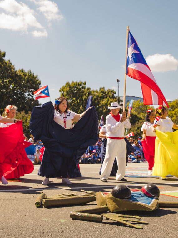 Dancers in flowing red, white and yellow costumes perform a traditional dance along the Benjamin Franklin Parkway as a participant carries a large Puerto Rican flag overhead.