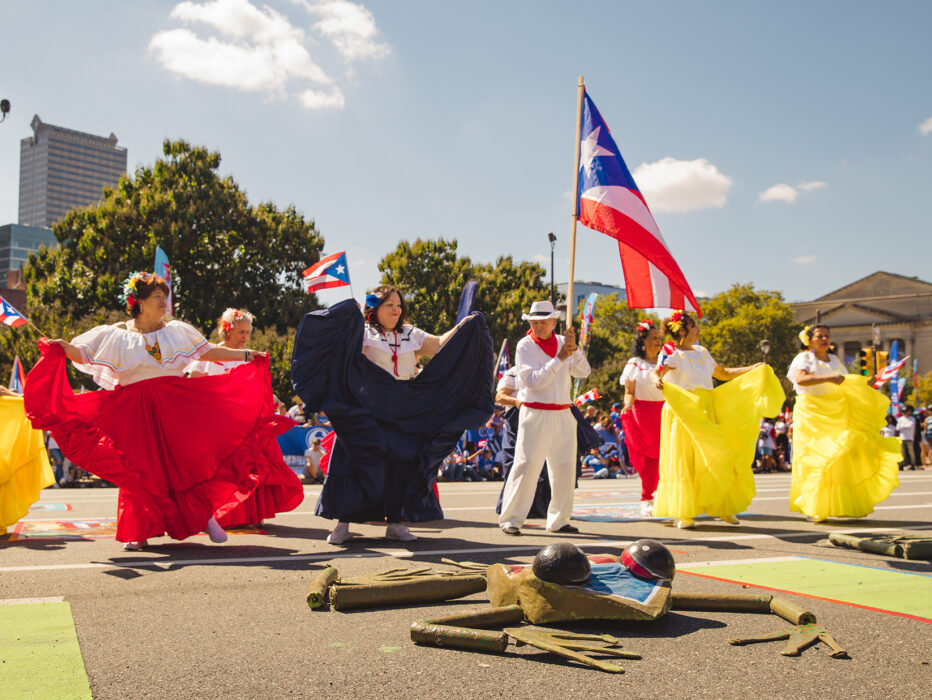 Des danseurs vêtus de costumes virevoltants rouges, blancs et jaunes exécutent une danse traditionnelle le long du Benjamin Franklin Parkway, tandis qu'un participant brandit au-dessus de sa tête un grand drapeau portoricain.