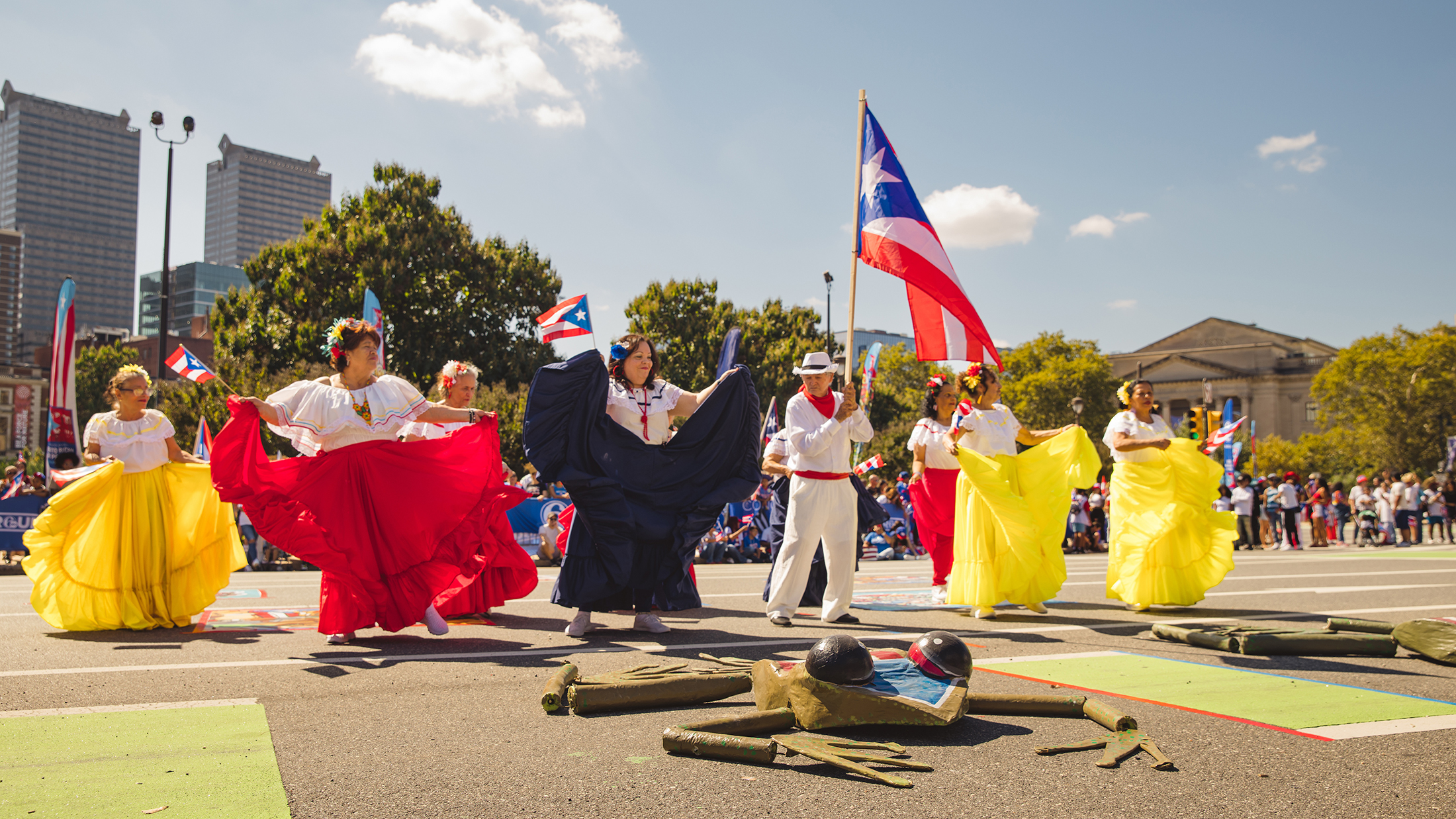Dancers in flowing red, white and yellow costumes perform a traditional dance along the Benjamin Franklin Parkway as a participant carries a large Puerto Rican flag overhead.