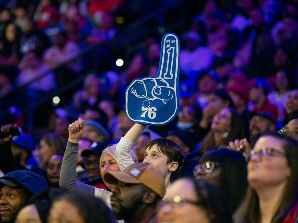 A young Philadelphia 76er fan holds a large blue foam finger reading "We're #1" above a packed crowd during game.