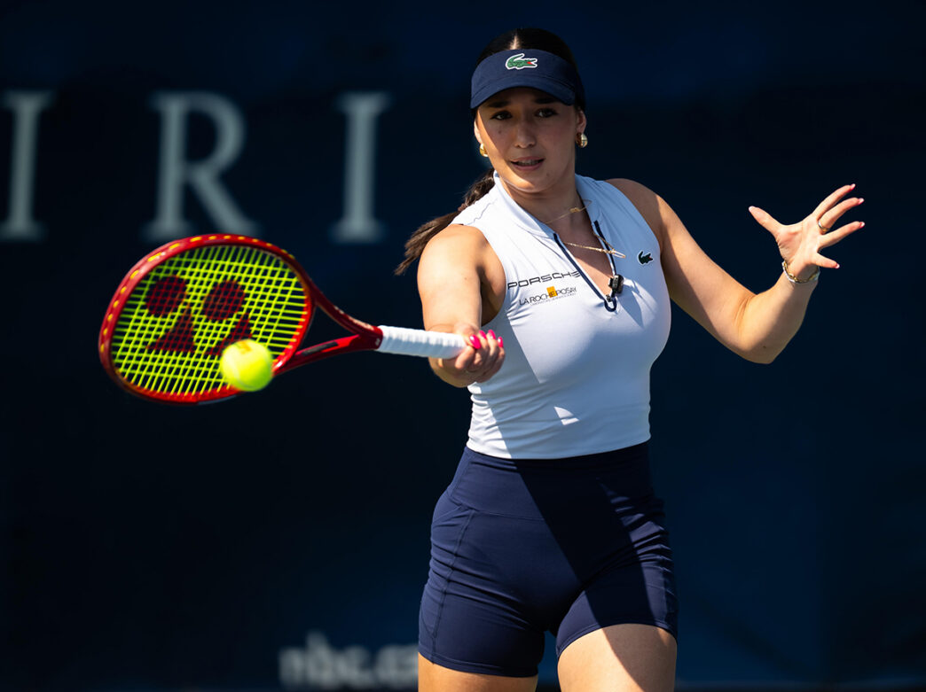 A tennis player reaches forward with a racquet to return a ball during a match, while wearing a shite sleeveless top, navy shorts and a visor.