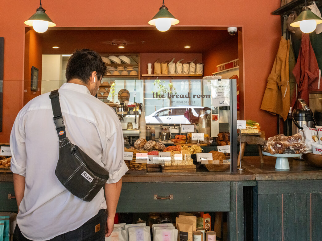 A person stands up at the counter of a bakery, with their back to the camera, looking through a glass partition at various breads and baked goods.
