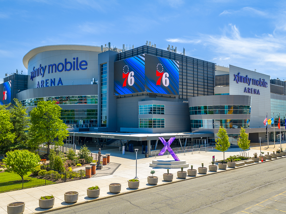 L'extérieur de la Xfinity Mobile Arena par une journée ensoleillée, avec de grands logos des Philadelphia 76ers affichés sur des écrans numériques au-dessus d'une entrée de la salle.