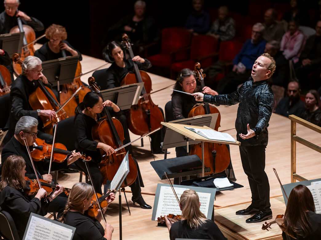 A conductor leads musicians of the Philadelphia Orchestra, including a section of cellists and violinists, during a performance before a seated audience.
