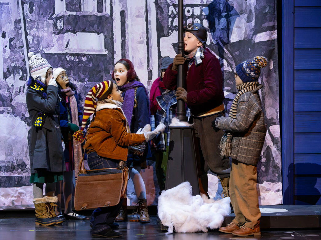 Young performers dressed in winter coats, hats and scarves gather around a lamppost during a production of A Christmas Story.