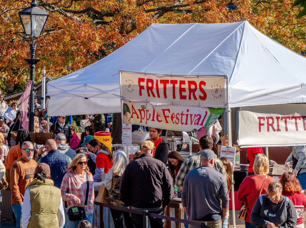 Festivalgoers lined up beneath a large white vender tent selling apple fritters during Peddler's Village Apple Festival, surrounded by vibrant autumn foliage.