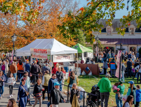Crowds walk along paved pathways through colorful fall foliage at Peddler's Village during the Apple Festival, with vendor tents selling apple fritters and more.