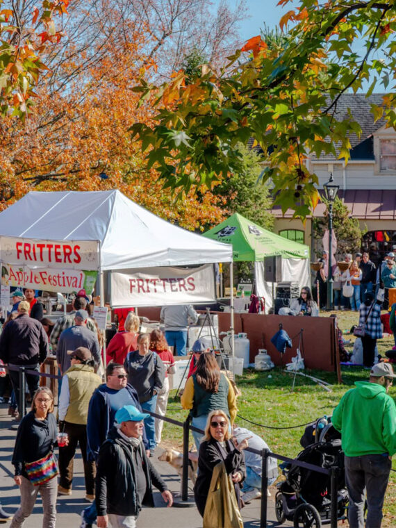 Crowds walk along paved pathways through colorful fall foliage at Peddler's Village during the Apple Festival, with vendor tents selling apple fritters and more.