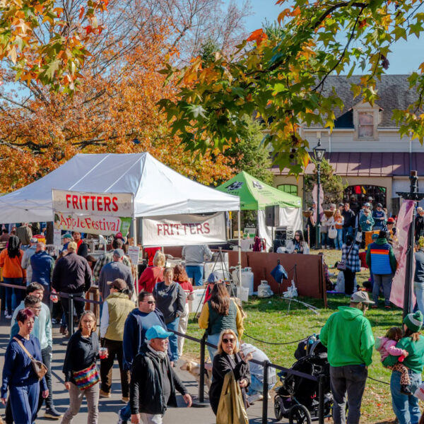 Crowds walk along paved pathways through colorful fall foliage at Peddler's Village during the Apple Festival, with vendor tents selling apple fritters and more.