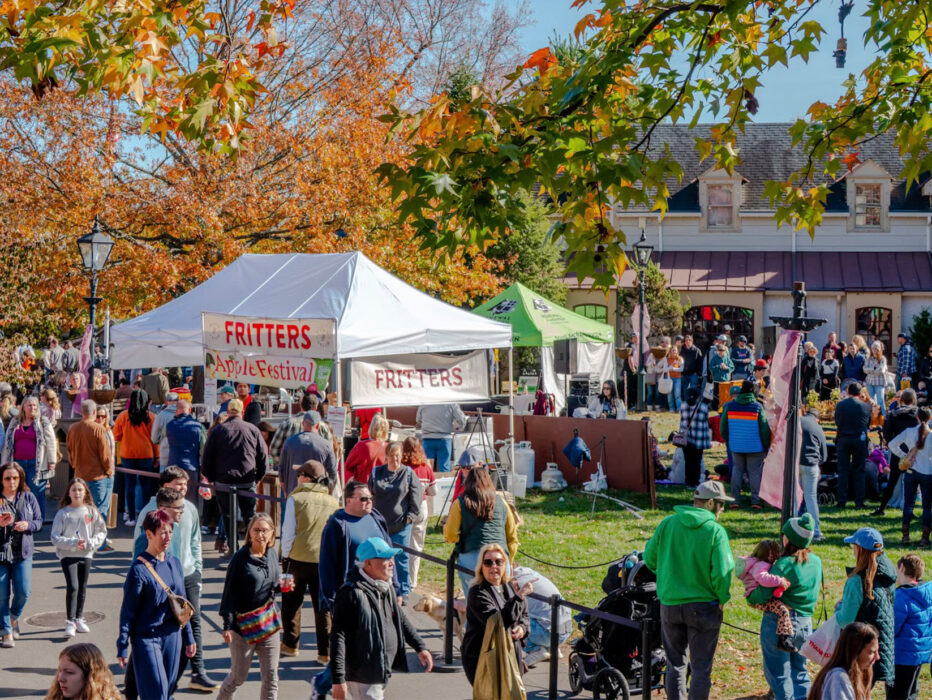 Crowds walk along paved pathways through colorful fall foliage at Peddler's Village during the Apple Festival, with vendor tents selling apple fritters and more.