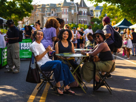 Trois personnes sont assises à une table au milieu d'une rue piétonne ; elles mangent et sourient tandis que les visiteurs flânent parmi les stands des exposants lors du « Dollar Stroll » de Baltimore Avenue.