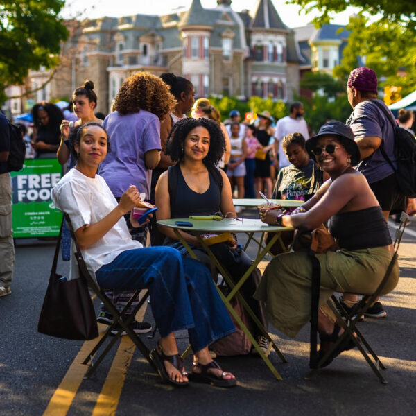 Three people sit at a table in the middle of a car-free street eating and smiling as visitors browse vendor tents during the Baltimore Avenue Dollar Stroll.