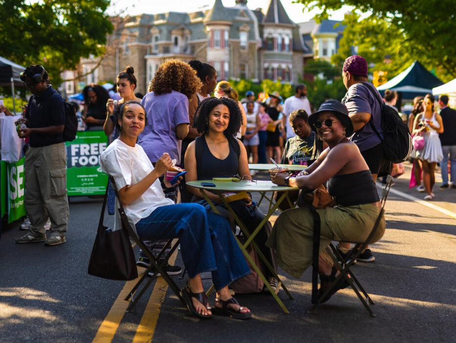 Trois personnes sont assises à une table au milieu d'une rue piétonne ; elles mangent et sourient tandis que les visiteurs flânent parmi les stands des exposants lors du « Dollar Stroll » de Baltimore Avenue.