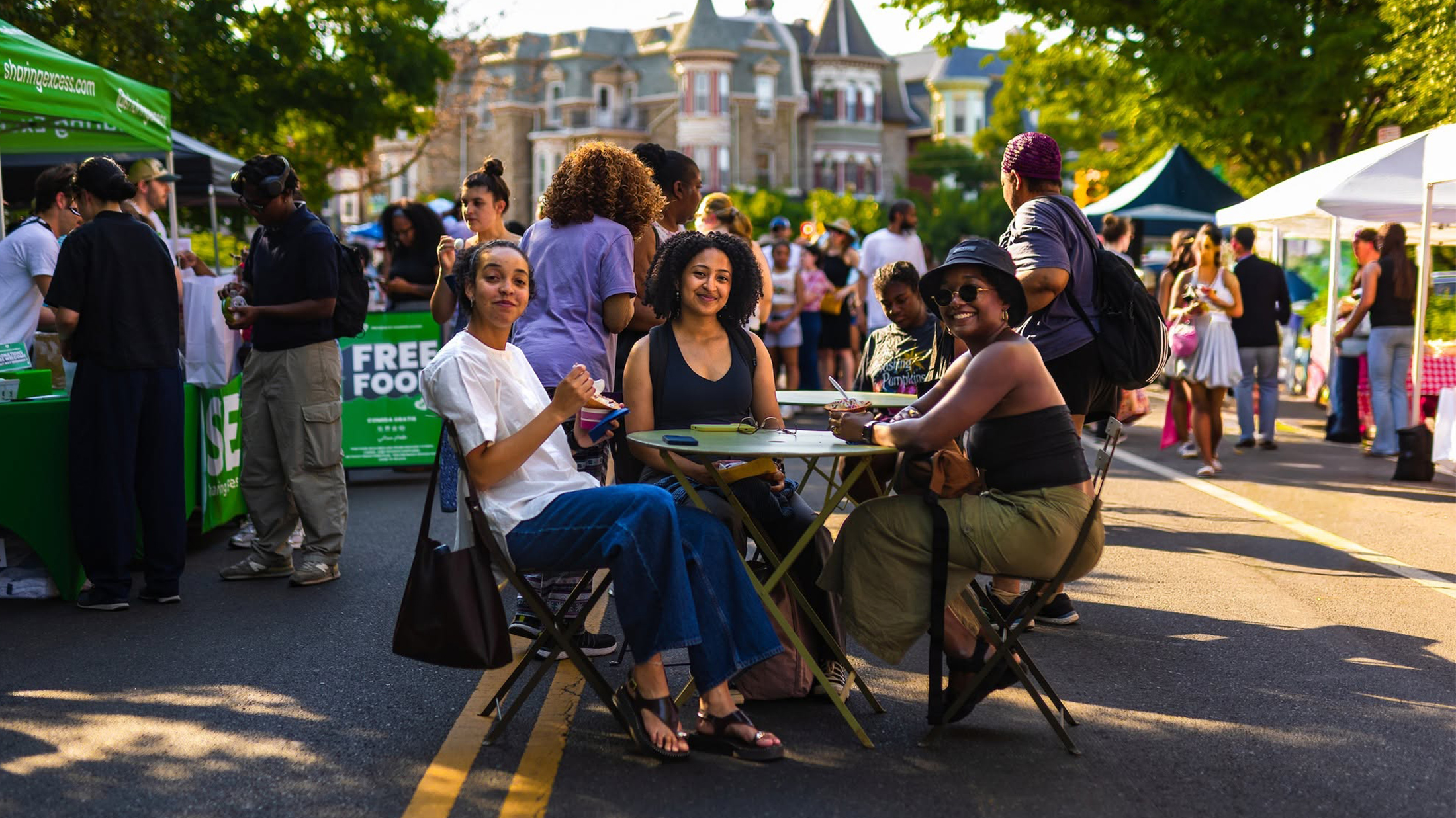 Trois personnes sont assises à une table au milieu d'une rue piétonne, en train de manger et de sourire, tandis que les visiteurs flânent parmi les stands des exposants lors du « Dollar Stroll » de Baltimore Avenue.
