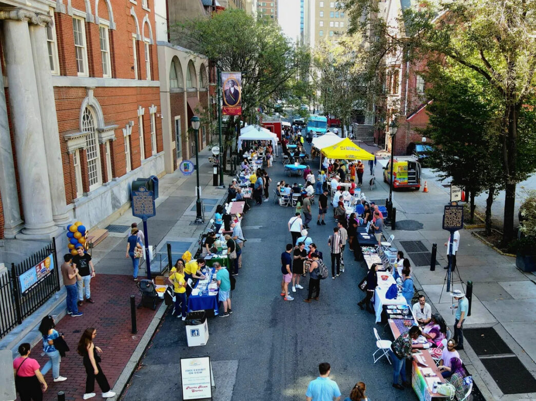 An overhead view of a block party showing visitors browsing tables, tents and community booths lining a street outside the brick building of the Historical Society of Pennsylvania.