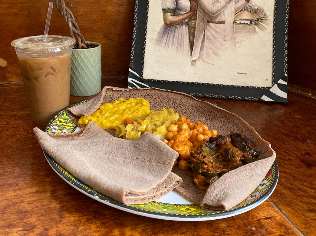 An Ethiopian breakfast platter with injera surrounds a serving of scrambled eggs, stewed cabbage, chickpeas and greens on a colorful plate, with an iced coffee, a potted plant and framed artwork on the tabletop in the background.
