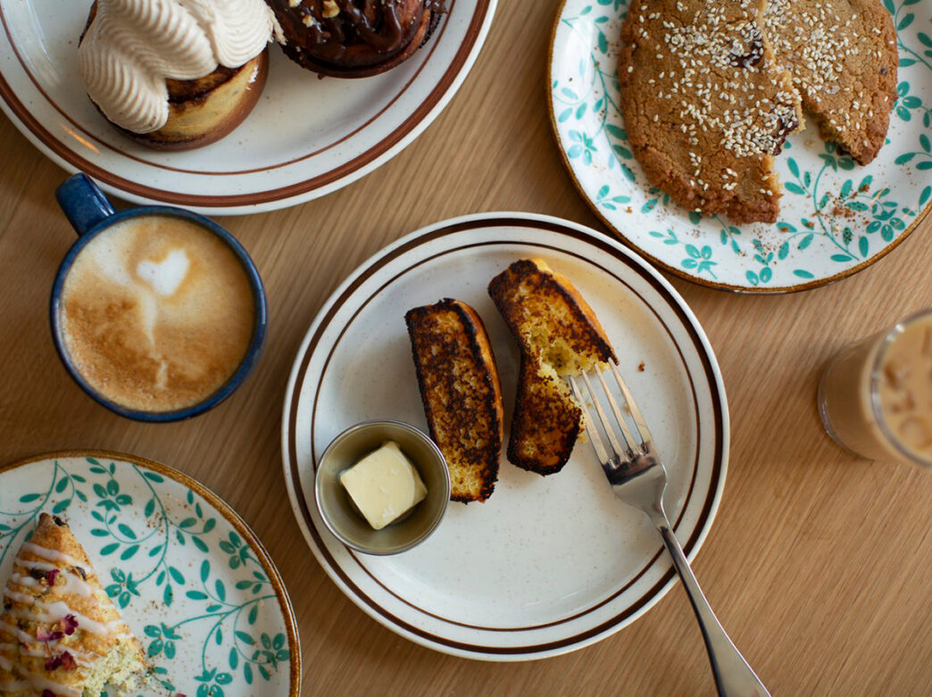 An overhead view of a cafe table with toasted bread and butter, pastries, sesame-topped cookie, a latte and an iced drink.