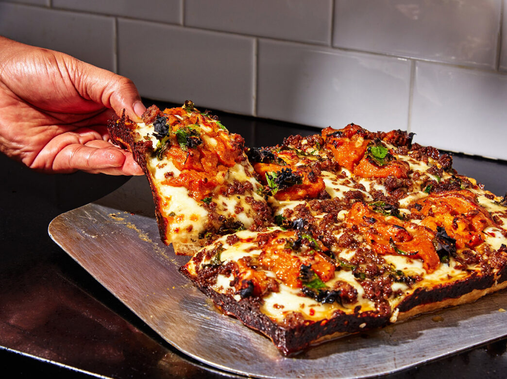A hand lifts a thick Detroit-style pizza slice topped with melted cheese, tomatoes and herbs from a freshly baked pie.