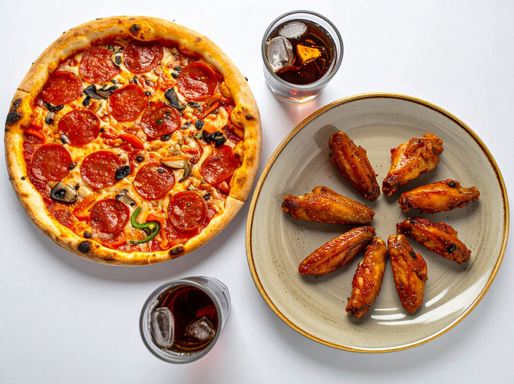 An overhead view of a pepperoni, mushroom and pepper pizza beside a plate of glazed chicken wings, with two glasses of iced soda on a white table.