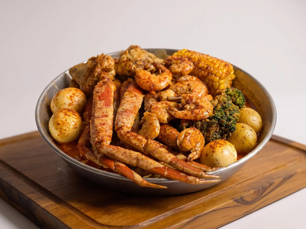 A large bowl of seasoned crab legs, shrimp, corn, potatoes and greens sits on a wooden serving board.