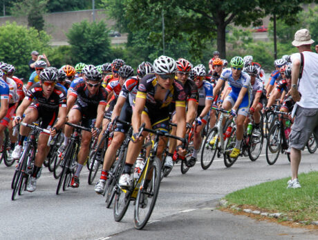 A large pack of competitive cyclists round a bend during a road race as a photographer captures the action from the roadside.