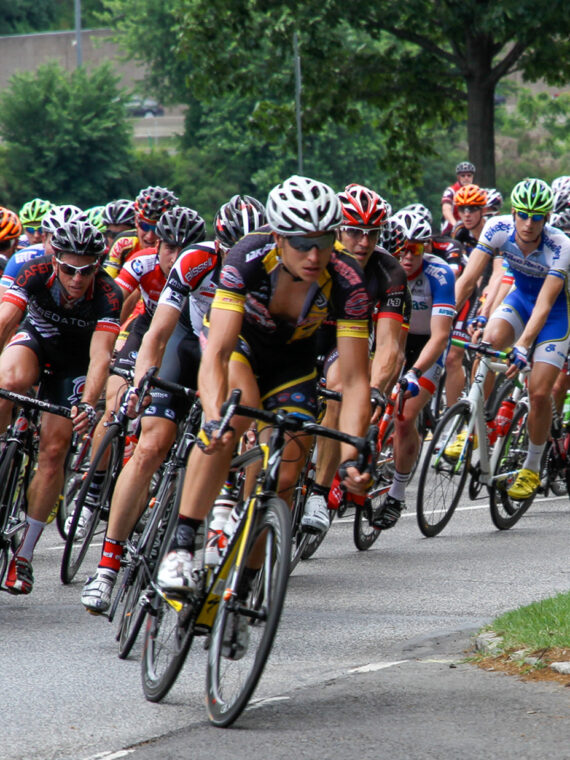 A large pack of competitive cyclists round a bend during a road race as a photographer captures the action from the roadside.