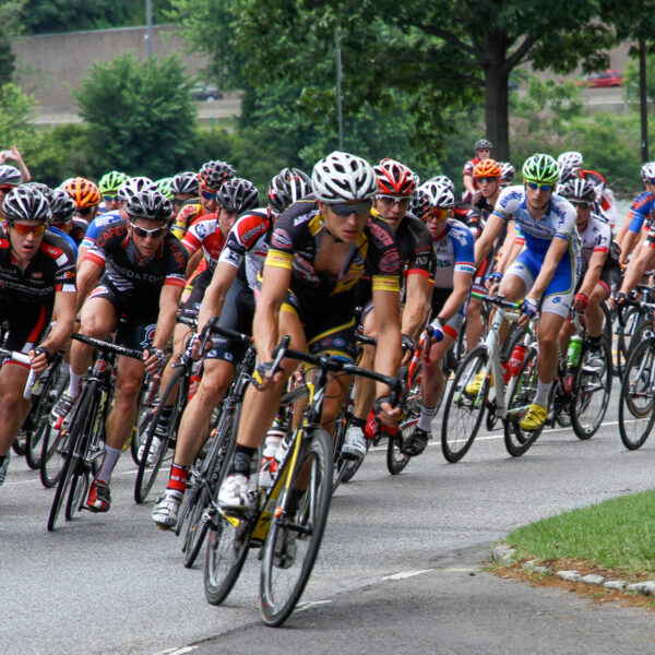 Een grote groep wedstrijdfietsers rijdt tijdens een wegwedstrijd een bocht om, terwijl een fotograaf de actie vanaf de kant vastlegt.