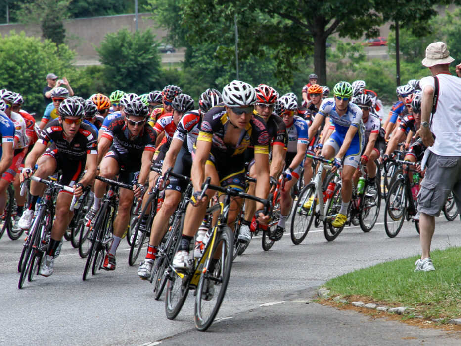 A large pack of competitive cyclists round a bend during a road race as a photographer captures the action from the roadside.