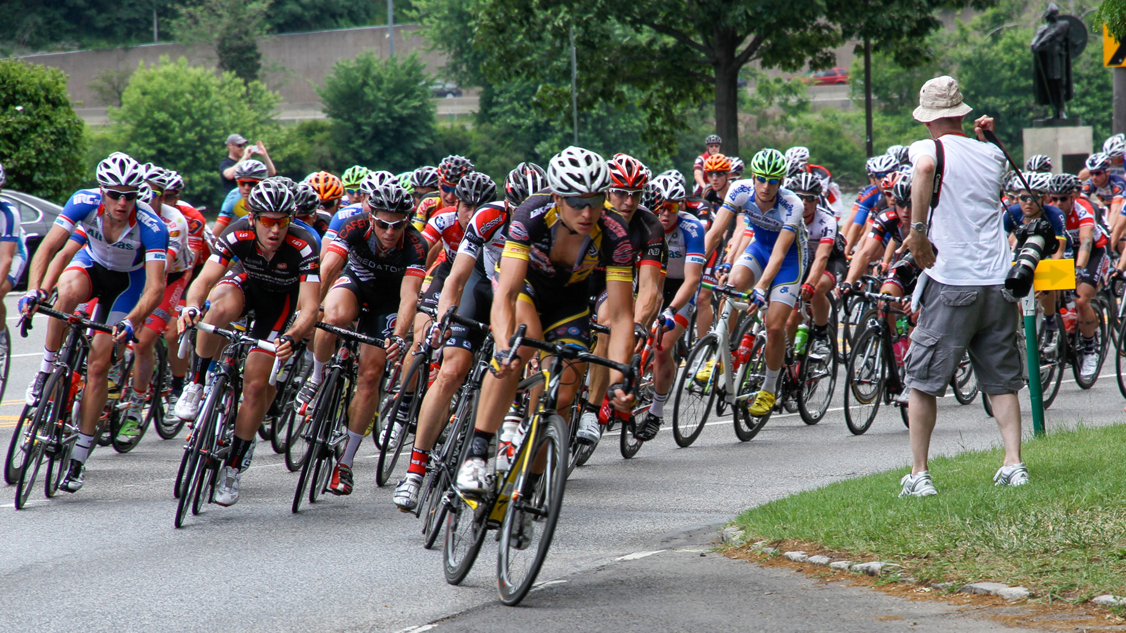 Un grand peloton de cyclistes de compétition négocie un virage lors d'une course sur route, tandis qu'un photographe immortalise la scène depuis le bord de la route.