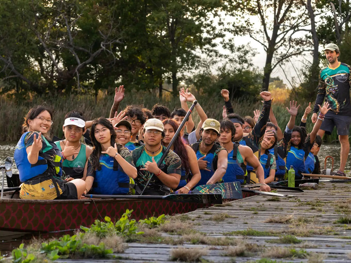 A group of paddlers wearing life jackets pose together in a dragon boat, smiling, waving and raising their hands beside a dock on a tree-lined waterway.