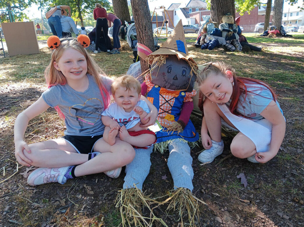 Three children pose with a straw scarecrow sitting on the ground at the Perkasie Fall Festival, with hay, trees and additional scarecrows int he background.