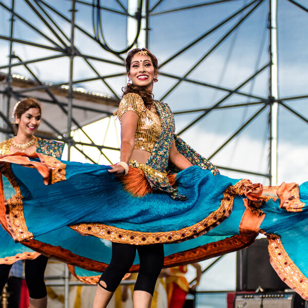 Tres bailarinas, con trajes vaporosos de color turquesa y naranja, dan vueltas y interpretan una danza tradicional india en un escenario al aire libre durante el Festival de la India de Filadelfia.