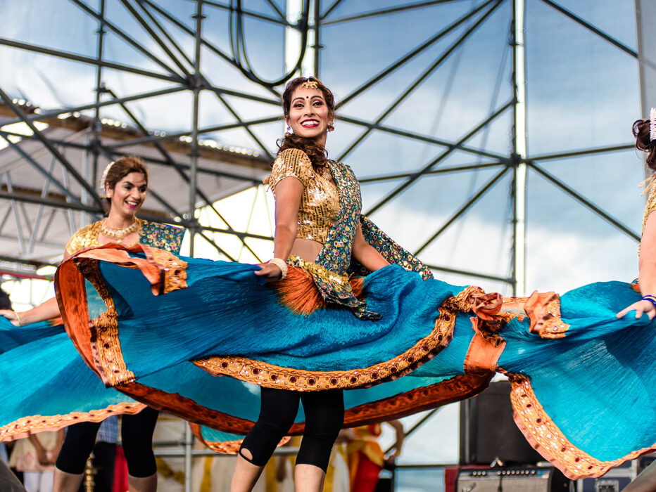 Three dancers in flowing turquoise and orange costumes twirl and perform a traditional Indian dance on an outdoor stage during Philadelphia's Festival of India.