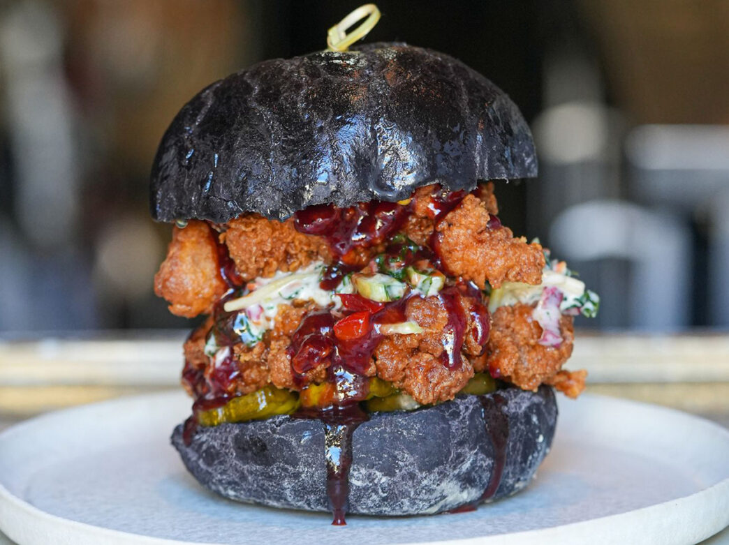 A towering fried chicken sandwich on a black charcoal bun is topped with creamy slaw, pickles and a barbecue-style sauce that drips down the sides of the sandwich.