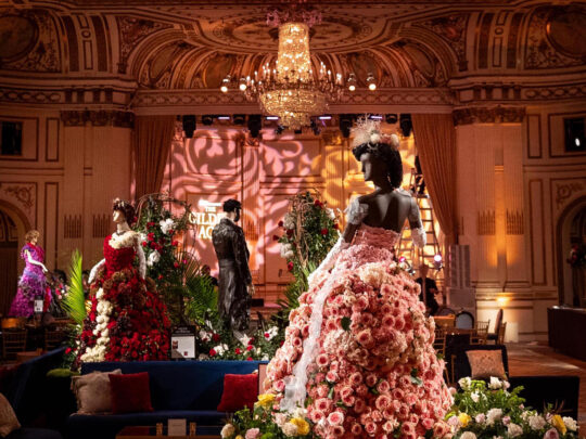 Elaborate mannequins dressed in gowns made from colorful flowers fill an ornate ballroom beneath a grand crystal chandelier, including a sweeping pink rose-covered dress in the center.