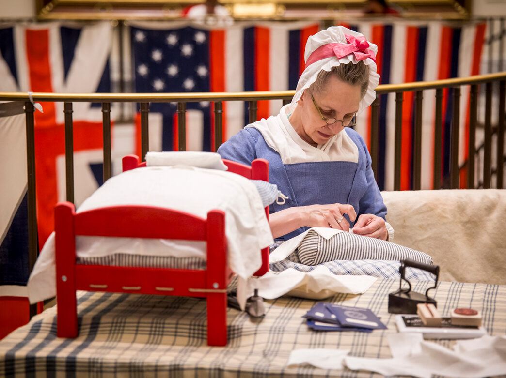 Een historische vertolker in kleding uit het koloniale tijdperk naait met de hand stof aan een tafel in een tentoonstelling in het Museum of the American Revolution.