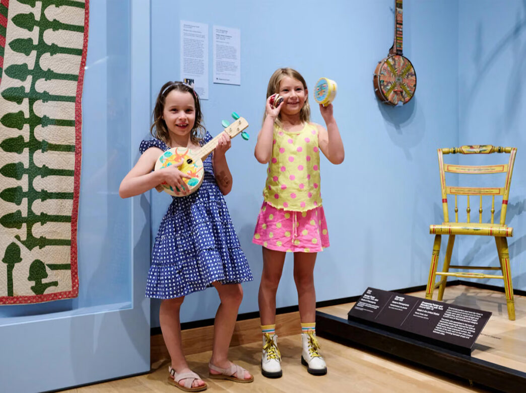 Two children smile while holding and playing colorful handmade instruments in a gallery the Philadelphia Museum of Art, surrounded by colorful decorative objects on display.