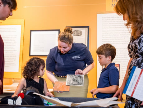 A museum educator wearing a navy polo shows historical materials to two children as their family gathers around an interactive exhibit.