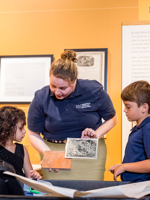 A museum educator wearing a navy polo shows historical materials to two children as their family gathers around an interactive exhibit.