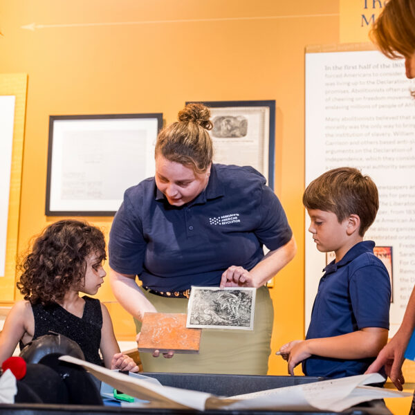 Un educador de museo, vestido con un polo azul marino, muestra materiales históricos a dos niños mientras su familia se reúne alrededor de una exposición interactiva.