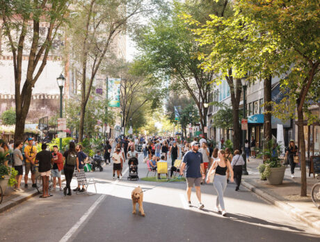Crowds stroll, gather and relax along a car-free, tree-lined Walnut Street during Open Streets: West Walnut, with outdoor seating, greenery, shops and restaurants lining the route.