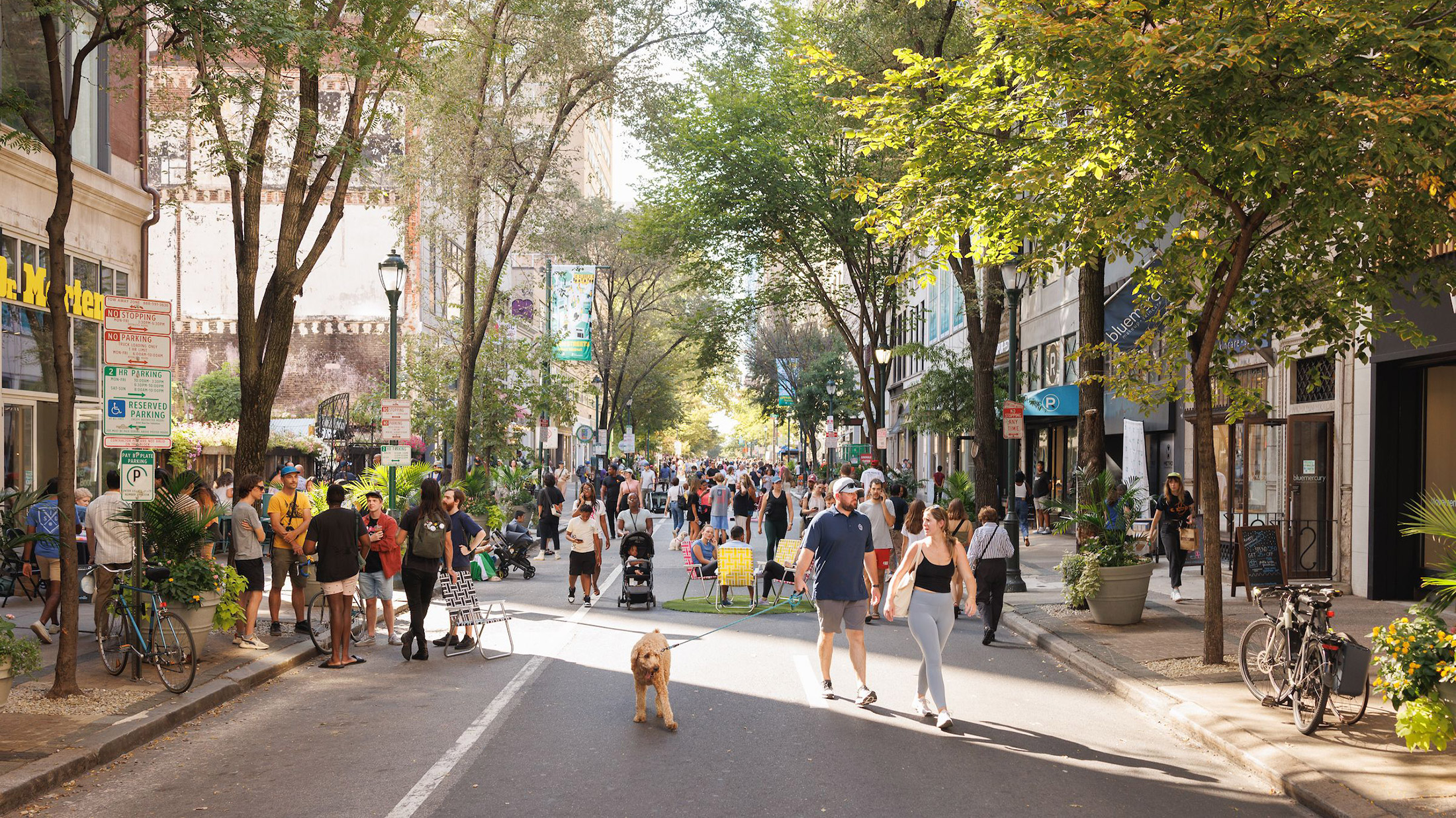 Crowds stroll, gather and relax along a car-free, tree-lined Walnut Street during Open Streets: West Walnut, with outdoor seating, greenery, shops and restaurants lining the route.