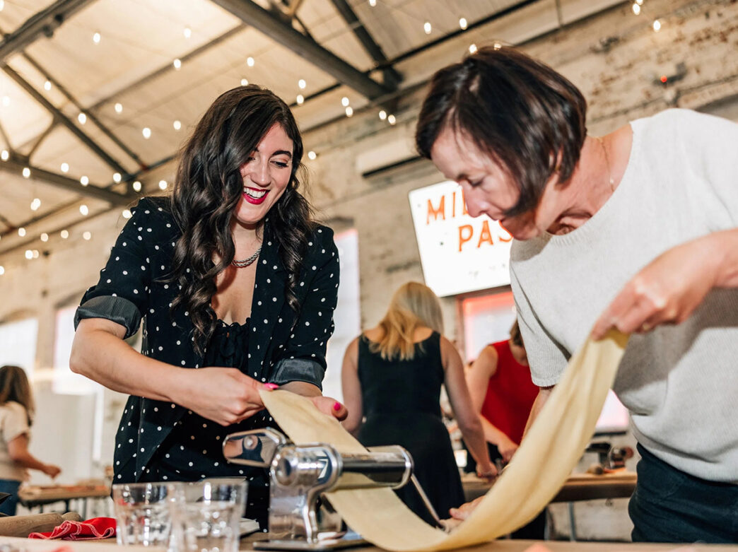 Two women smile as they feed long sheets of fresh pasta dough through a hand-cranked pasta machine during a group cooking class.
