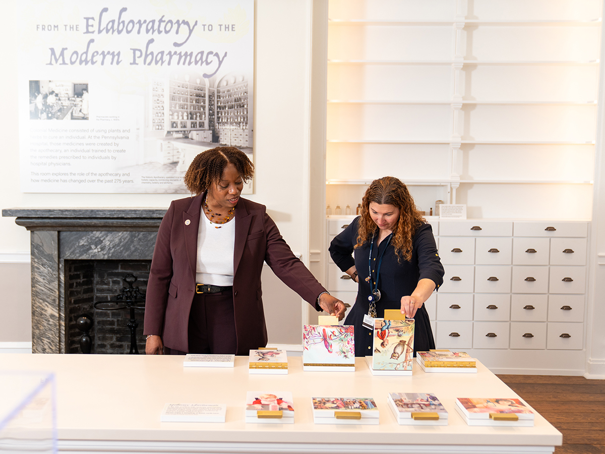 Two people examine an interactive display table in a museum exhibit exploring the evolution of pharmacy, with historic white cabinetry and black fireplace in the background.