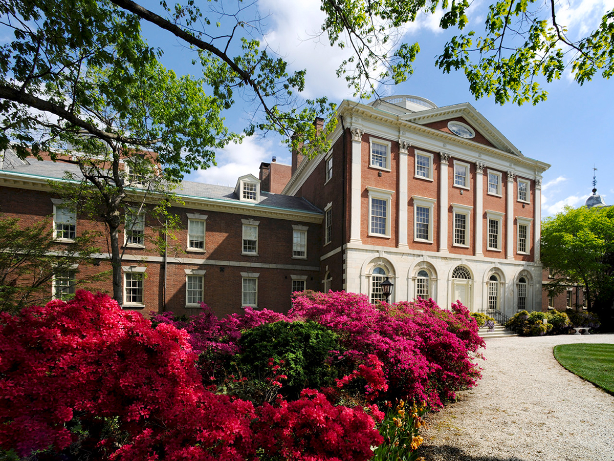 The historic red-brick Pennsylvania Hospital building framed by leafy green trees and bright pink flowering shrubs on a sunny day.